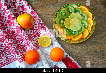 Sobre la mesa es un plato de rodajas de frutas: naranjas, kiwi, limón. Cerca de ellos en una hermosa toalla son dos naranjas y una manzana. Foto de stock