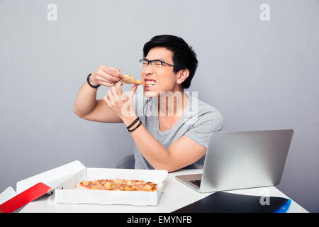 Apuesto hombre asiático sentado a la mesa y comer pizza sobre fondo gris Foto de stock