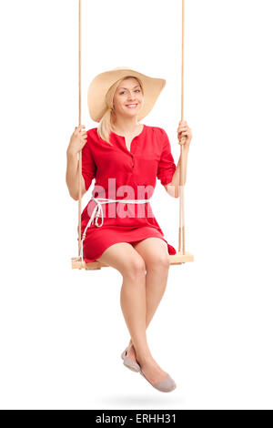 Bella mujer en un vestido rojo y un elegante sombrero balanceándose en un columpio de madera aislado sobre fondo blanco. Foto de stock