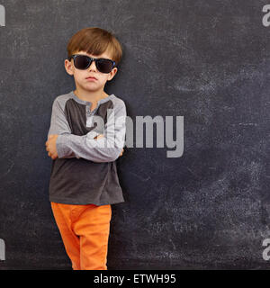 Retrato de niño inteligente con gafas de sol de pie con sus manos contra la pizarra con copyspace plegada.