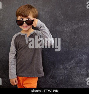 Retrato de un niñito en gafas de sol mirar lejos en el espacio de copia. Cool boy mirando por encima de sus gafas de sol contra la pizarra.