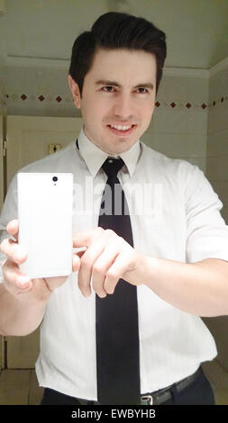 Hombre de negocios sonriendo feliz teniendo selfie. Joven modelo masculino en suite haciendo una foto de él en el cuarto de baño delante del espejo. Concepto Foto de stock