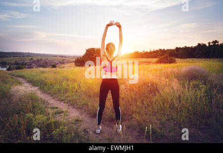 Joven mujer deportiva con los brazos levantados durante la puesta de sol en verano