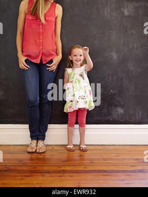 Imagen de cute little girl de pie con su madre en la casa. Madre e hija juntos contra una pared negra, adentro.