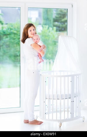 La joven madre de llevar a su bebé a dormir en una cama con dosel blanco junto a una gran ventana Foto de stock