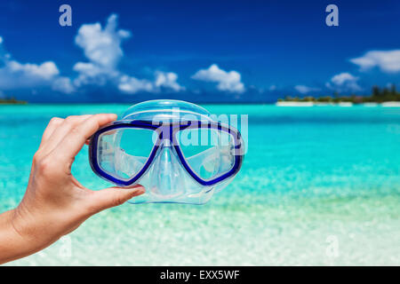Mano sujetando snorkel googles contra playa borrosa y cielo azul