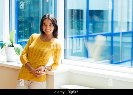 Sonriente atractiva empresaria de pie cerca de la ventana en la oficina y mirando a la cámara