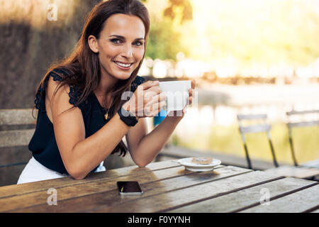 Retrato de la hermosa joven sentado en una mesa con una taza de café en la mano mirando a la cámara sonriendo mientras en el cafe.
