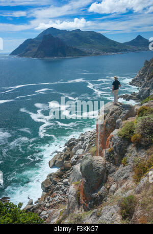 Un caminante solitario parado sobre roca encima del Océano Atlántico mira hacia Pico centinela seascape, visto desde Chapman's Peak Drive Foto de stock