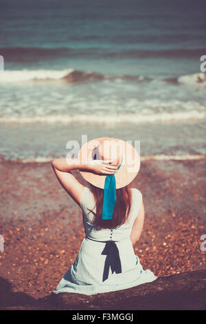 Joven y bella muchacha sentada histórico en la playa en un vestido azul y llevaba un sombrero de paja