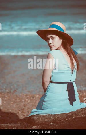 Joven y bella muchacha sentada histórico en la playa en un vestido azul y llevaba un sombrero de paja