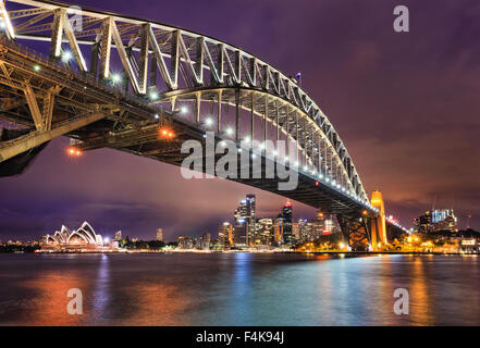 East Side del Sydney Harbour Bridge al atardecer con iluminación brillante de arco de acero y columnas reflejando en las aguas borrosa