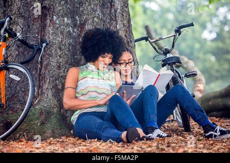 Mujer madura e hijo sentarse contra el árbol del parque libro de lectura y tableta digital