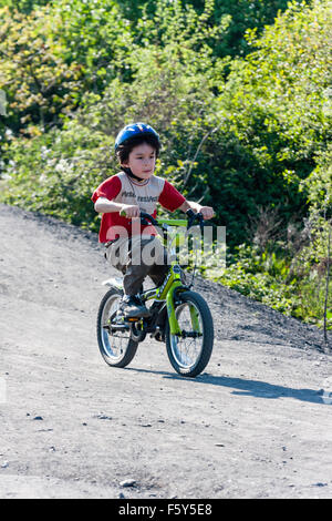 Niño de 9 años caucásica, boy, montando en bici verde pendiente abajo dirt track con arbustos en segundo plano. Lleva casco azul. En verano, el sol. Foto de stock