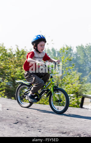 Niño de 9 años caucásica, boy, montando en bici verde pendiente abajo dirt track con arbustos en segundo plano. Lleva casco azul. En verano, el sol. Foto de stock