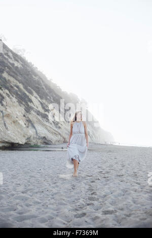 Mujer rubia caminando en una playa de arena, cerca de un acantilado.