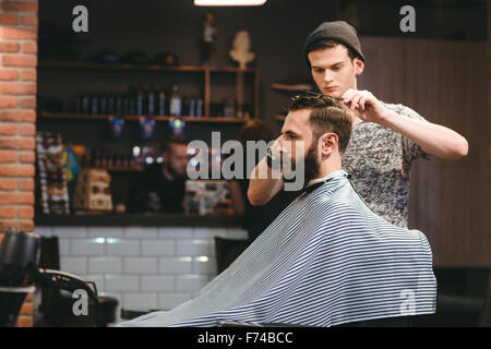 Joven apuesto barber hacer recorte de valoración del atractivo hombre barbado en el barbershop Foto de stock