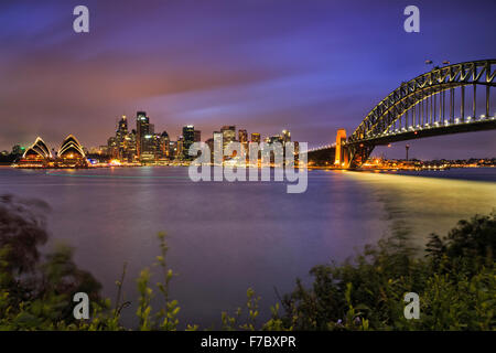 Los hitos de la ciudad de Sydney CDB cruzando el puerto con arco del puente, rascacielos y casa al atardecer con iluminación completa Foto de stock