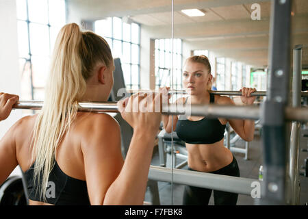 Mujer rubia caucásica muscular en un gimnasio haciendo flexiones con un barbell sobre sus hombros mientras se mira en el espejo. Mujer joven workin Foto de stock