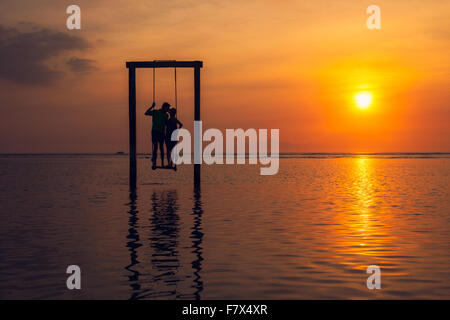 Silueta de un hombre y una mujer besándose, de pie en un columpio en el mar al atardecer, Indonesia
