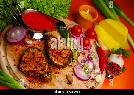 Cerca de la tabla de cortar de madera con dos lonchas de carne a la brasa con especias y verduras y queso alrededor y un tamiz Foto de stock