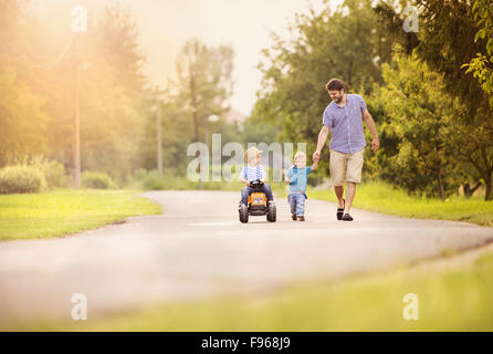 Feliz padre joven con sus dos pequeños hijos se divierten en la carretera, un niño es montar en el tractor pequeño, otro muchacho es la celebración del padre Foto de stock