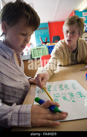 Chica sentada en una mesa en el aula practicar habilidades de escritura; con el profesor ayudando, Foto de stock