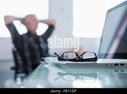 Los anteojos y el portátil en el escritorio en la oficina, el hombre relajante en segundo plano.