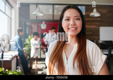 Sonriente joven empresaria mirando a la cámara y sus colegas están de pie en el fondo. Joven asiático femenino pro creativa