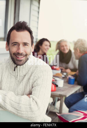 Retrato morena sonriente hombre de suéter en el porche con la familia en segundo plano.