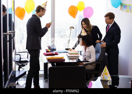  Mujer de negocios celebrando un cumpleaños y haciendo una fiesta con colegas en su oficina. Un amigo con teléfono móvil toma fotografías o Fotografía de stock