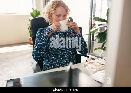 Retrato de mujer joven sentado en su pupitre bebiendo café y mirando el monitor del equipo. Diseñador reflexiva con café.