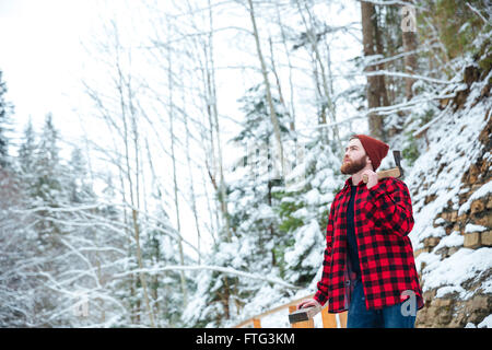 Grave joven barbudo en camisa a cuadros con ax en invierno bosque permanente Foto de stock
