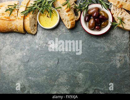 Ingredientes de comida italiana el fondo con rebanadas de pan ciabatta, aceite de oliva, las aceitunas y el romero en piedra gris pizarra