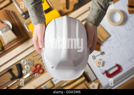 Manos sosteniendo un casco de seguridad protector contra una mesa de trabajo con hardware y herramientas de construcción, vista superior Foto de stock
