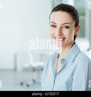 Sonriente joven y bella mujer de negocios sonriendo y mirando a la cámara, en el interior de la oficina del fondo