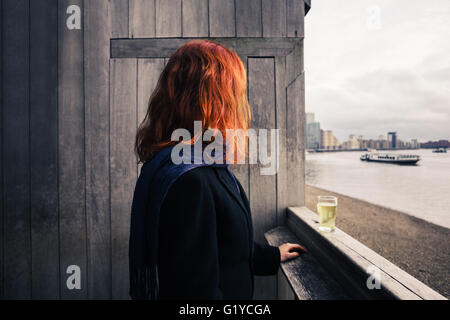 Una mujer joven está de pie sobre una terraza de madera por el río con un vaso de cerveza