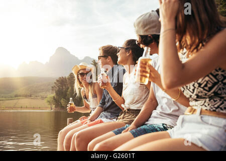 Retrato de los jóvenes, hombres y mujeres sentadas en jetty con cervezas. Los jóvenes hombres y mujeres disfrutando de un día en el lago.