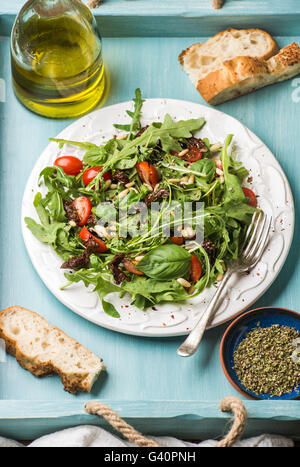 Con ensalada de rúcula, tomates cherry, piñones y hierbas en placa de cerámica blanca sobre fondo de madera azul, el enfoque selectivo Foto de stock