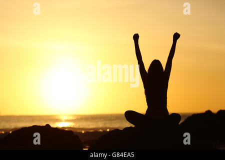 Vista posterior de un emocionado eufórica mujer silueta levantando los brazos y mirando al sol naranja en la playa al amanecer.