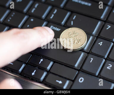 Una libra británica coin acostada sobre el teclado del portátil con el dedo pulsando la tecla Intro Foto de stock