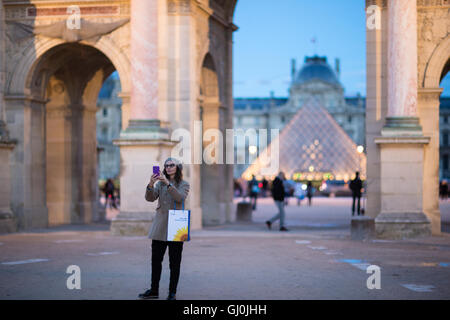 Una mujer que toma un selfie al Arc de triomphe du Carrousel & Palais du Louvre al atardecer, París, Francia