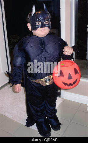 Un joven serio en un traje de Batman posee un plástico naranja calabaza mientras espera en la puerta a recoger dulces de sus vecinos en la víspera de Halloween en California, Estados Unidos. Foto de stock