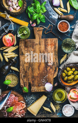 Ingredientes para cocinar la comida italiana sobre fondo oscuro con una tabla de cortar de madera rústica en el centro, vista superior, espacio de copia, vertical Foto de stock