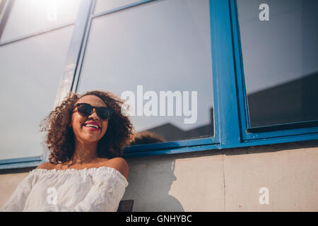 Disparo de feliz joven africana sentado afuera mirando lejos y sonriente. Mujer con gafas de sol sentado afuera mirando h Foto de stock