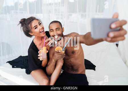 Sonriente, feliz pareja haciendo selfie multirracial y la celebración de cócteles en la playa.