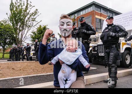 Un hombre vestido con un 'anónimo' de Guy Fawkes mask y sosteniendo un bebé, ofrece una desafiante salute delante de una fila de PSNI policías Foto de stock
