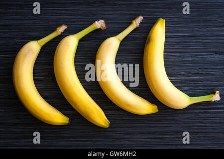 Cuatro plátanos maduros establecen en una fila sobre fondo oscuro de cerca Foto de stock