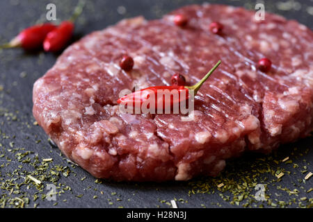 Primer plano de una hamburguesa cruda condimentada con pimienta roja, pimientos y hierbas en una superficie de pizarra Foto de stock