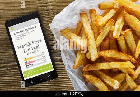 Contador de calorías concepto - teléfono móvil con contador de calorías app en la pantalla y papas fritas. Mesa de madera como fondo Foto de stock